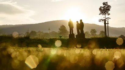 Drei Figuren auf Sockeln im Gegenlicht vor bergiger Landschaft bei Sonnenuntergang