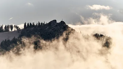 Berggipfel mit Kreuz umgeben von Nebel und Bäumen