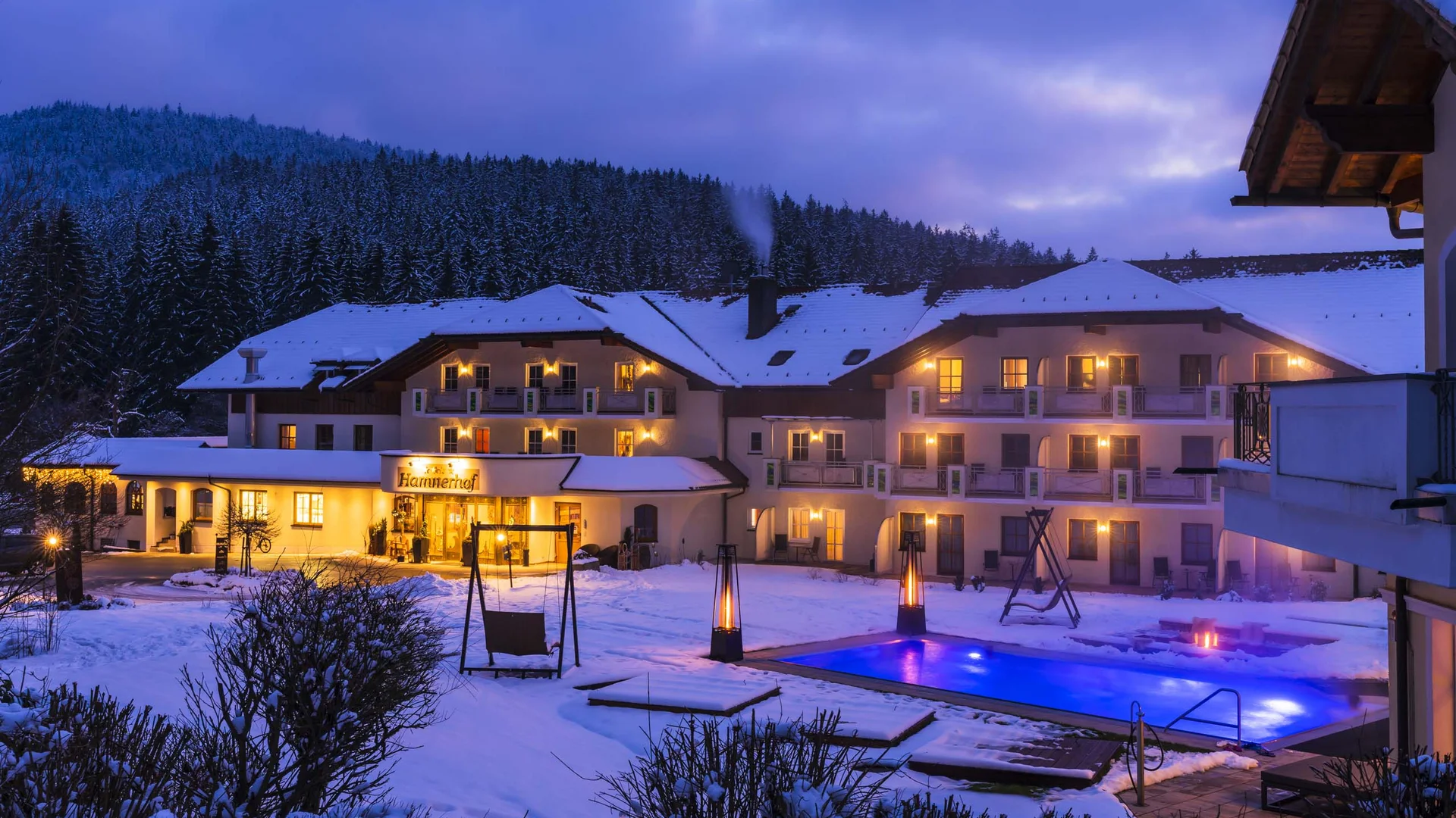 Verschneites Hotel mit beleuchtetem Pool und Wald im Hintergrund bei Abenddämmerung
